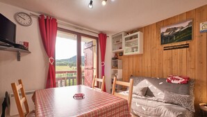 A cozy room with a wooden wall, a dining table with a checkered tablecloth, a sofa with a blanket, a clock, and a window with red curtains.