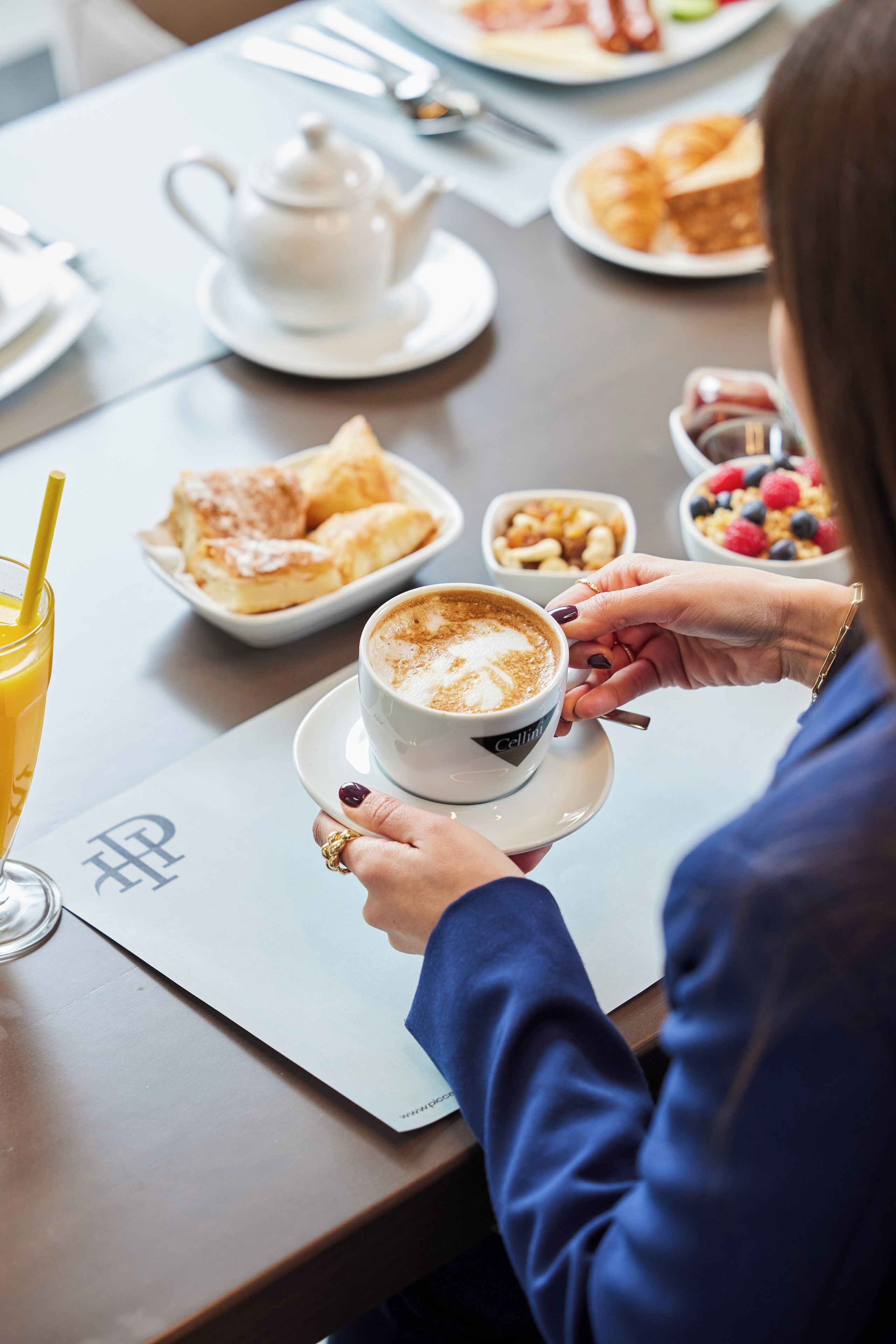 A person seated at a table with a cup of coffee, a teapot, and various food items.