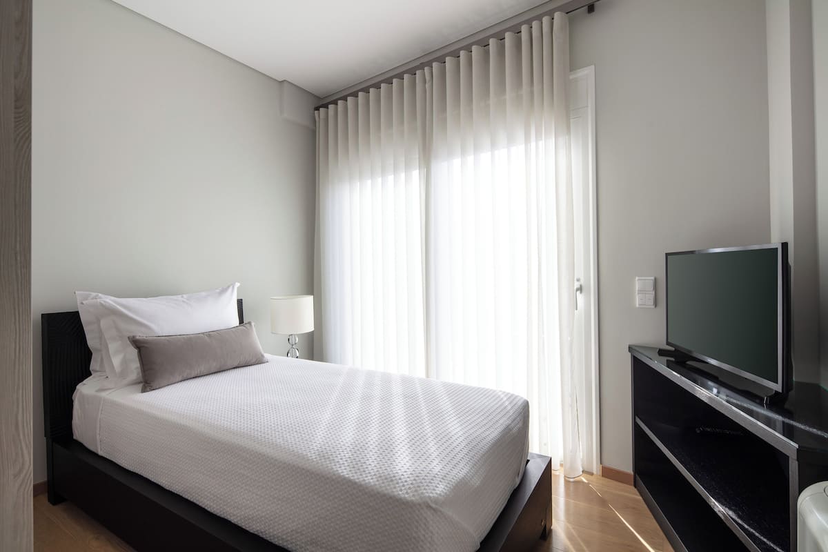 Une chambre d’hôtel moderne avec un lit soigneusement fait, un téléviseur à écran plat posé sur un meuble noir et une grande fenêtre ornée de rideaux légers.