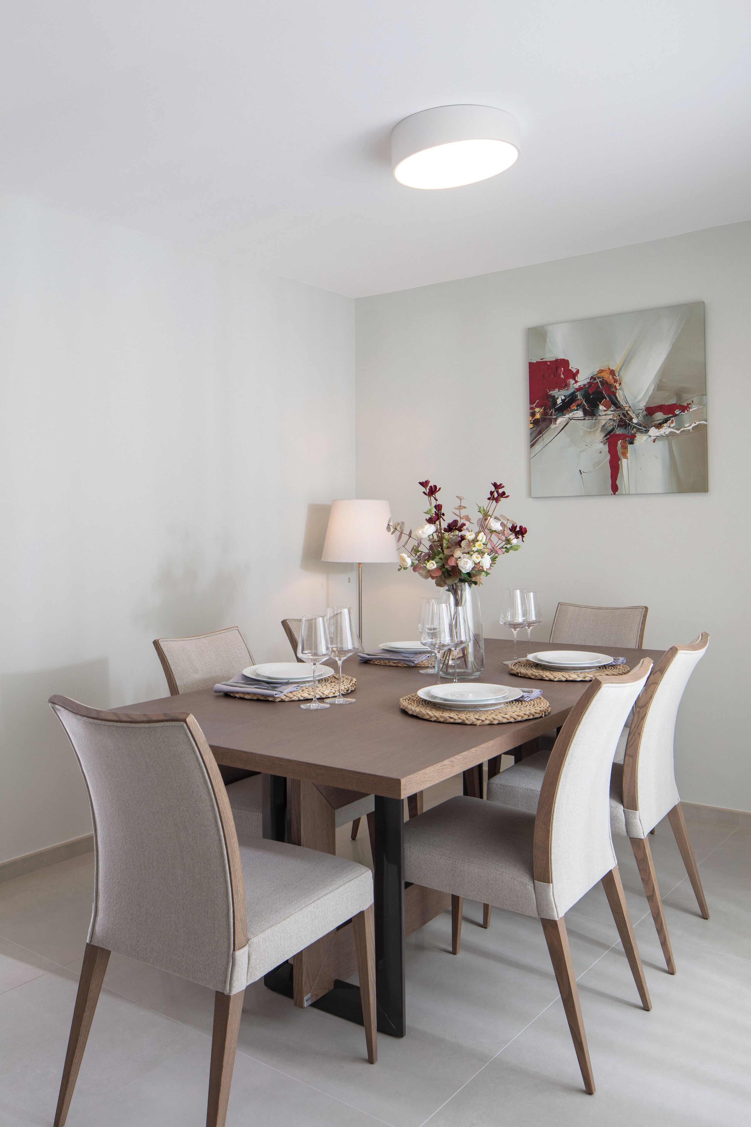 Un comedor con una mesa rectangular, seis sillas, un jarrón con flores y una lámpara de pared.