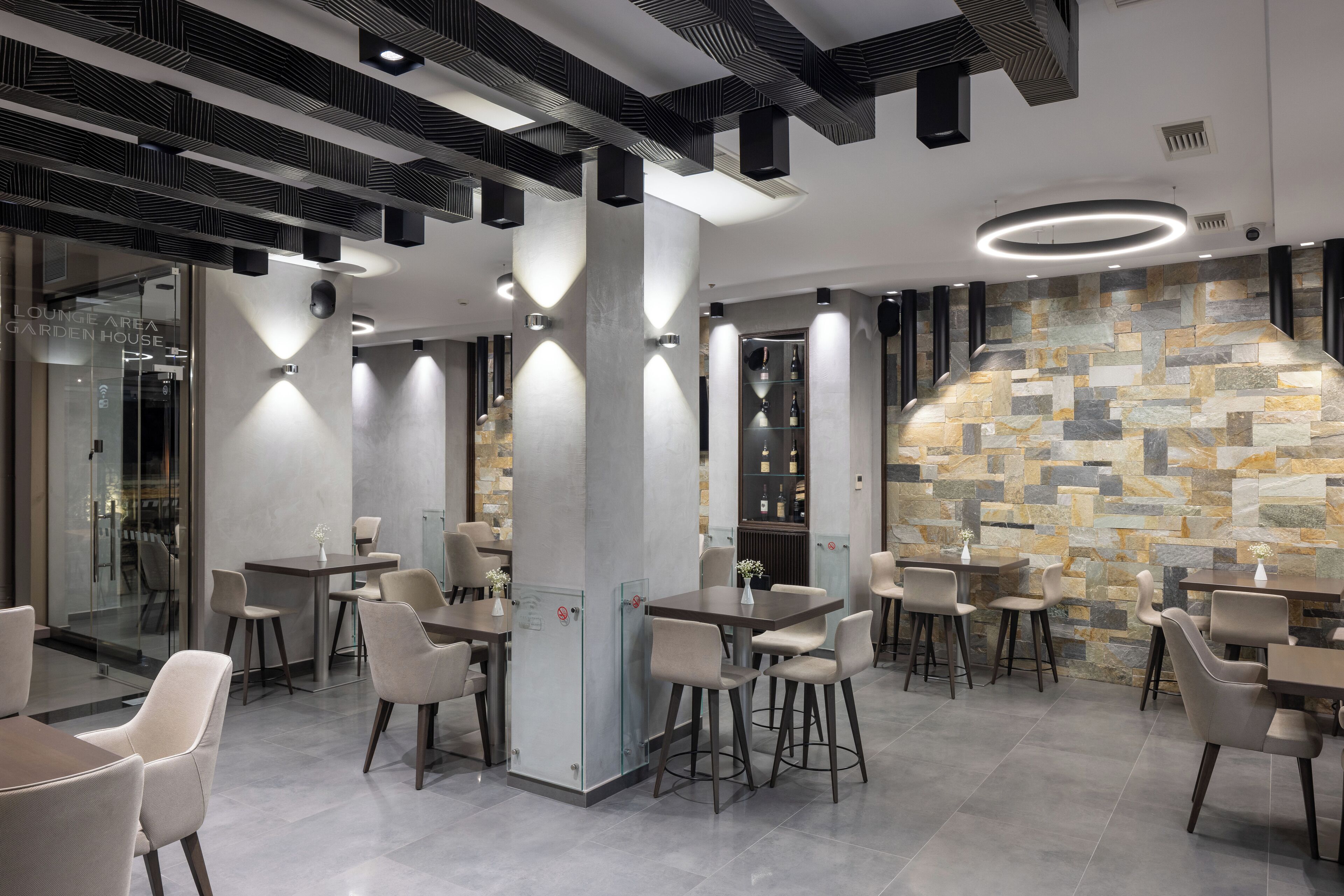 Een moderne zithoek met meerdere tafels en stoelen, een stenen muur en inbouwverlichting.