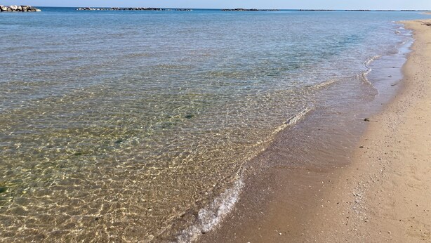 Una playa con aguas cristalinas y poco profundas, una orilla arenosa y afloramientos rocosos a lo lejos.