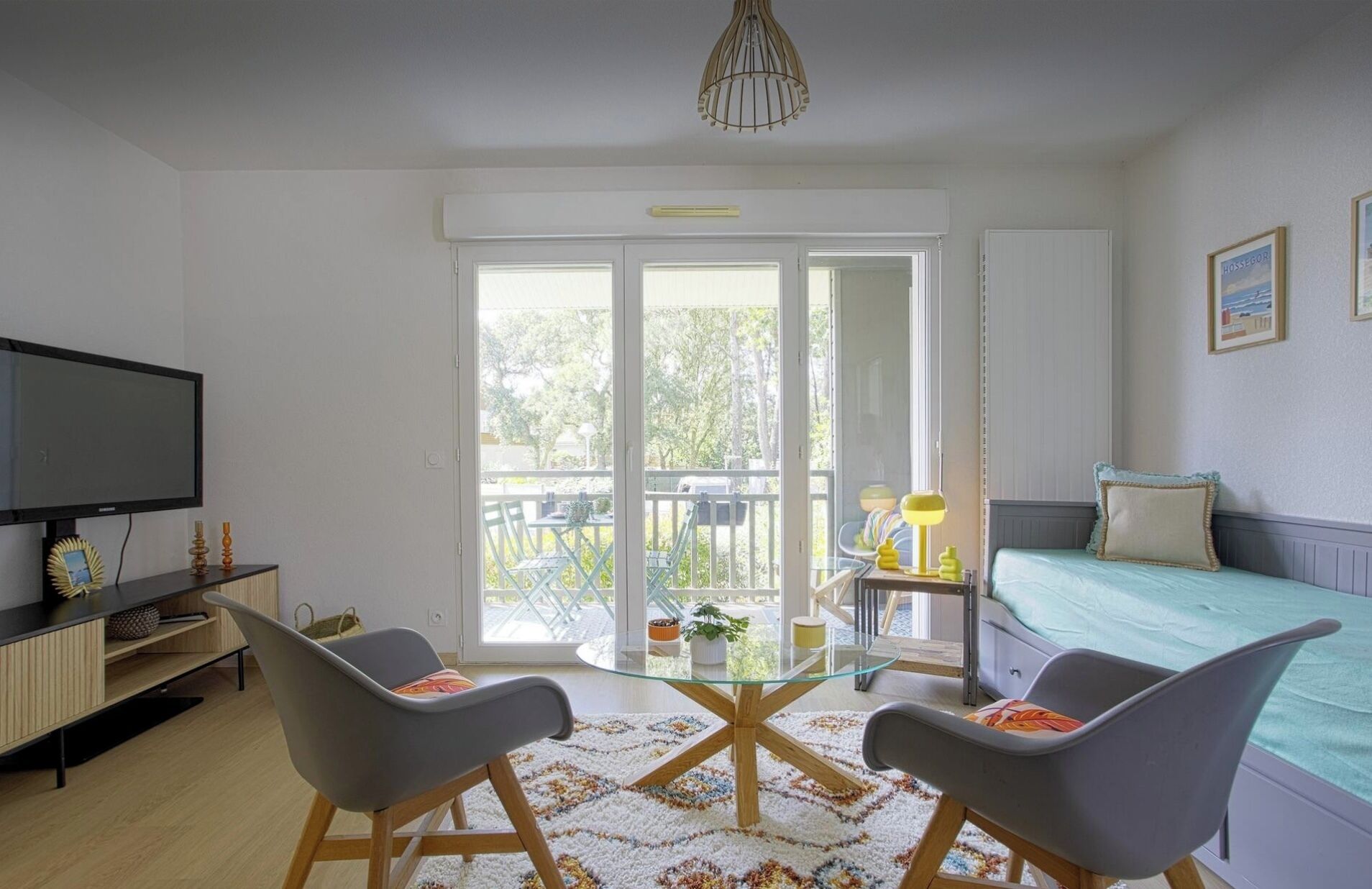 A modern living room with a glass-top coffee table, two grey chairs, a TV, and a bed with a blue bedspread.