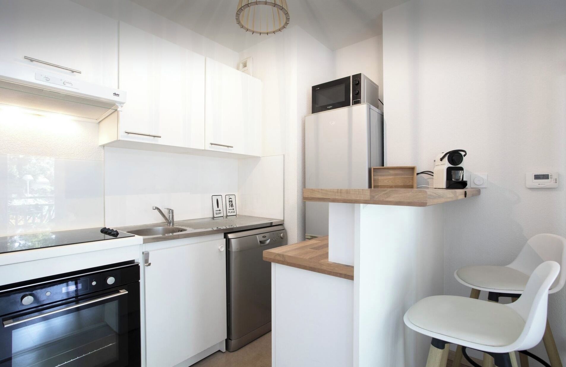 A compact kitchen with white cabinets, a built-in oven, a dishwasher, a microwave, and a coffee machine on a wooden shelf. There are two white chairs and a small table.