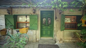 Een rustiek gebouw met een groene deur, twee ramen met groene luiken, een fiets en een vogelhuisje.