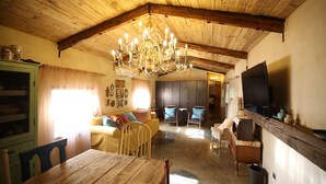 Een gezellige woonkamer met houten plafond, een kroonluchter, een aan de muur bevestigde televisie en een eettafel met stoelen.