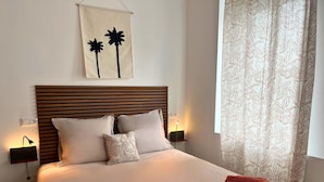 A neatly made bed with a wooden headboard, two bedside lamps, and a wall-mounted artwork of palm trees. - Apart'Moment EN GROUPE - L'Horizon Bleu (Bourbourg)