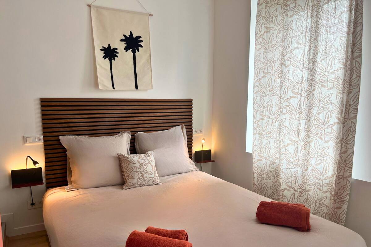 A neatly made bed with a wooden headboard, two bedside lamps, and a wall-mounted artwork of palm trees.