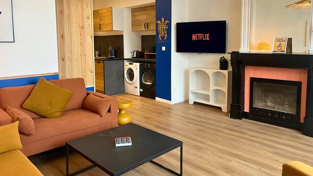 A modern living room with a sofa, coffee table, and a flat-screen TV. A kitchen is visible in the background with a washing machine. - Apart'Moment EN GROUPE - L'Horizon Bleu (Bourbourg)