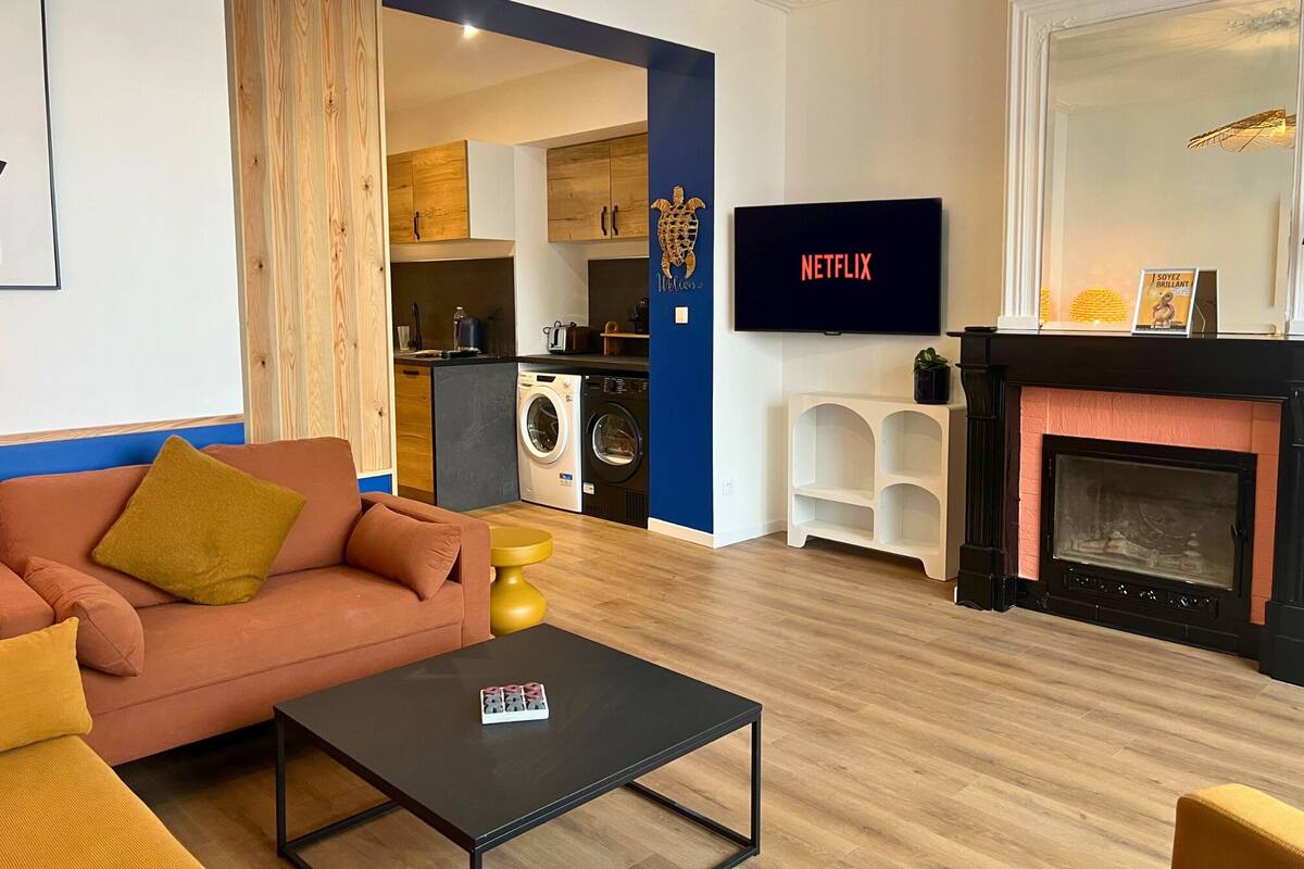 A modern living room with a sofa, coffee table, and a flat-screen TV. A kitchen is visible in the background with a washing machine.