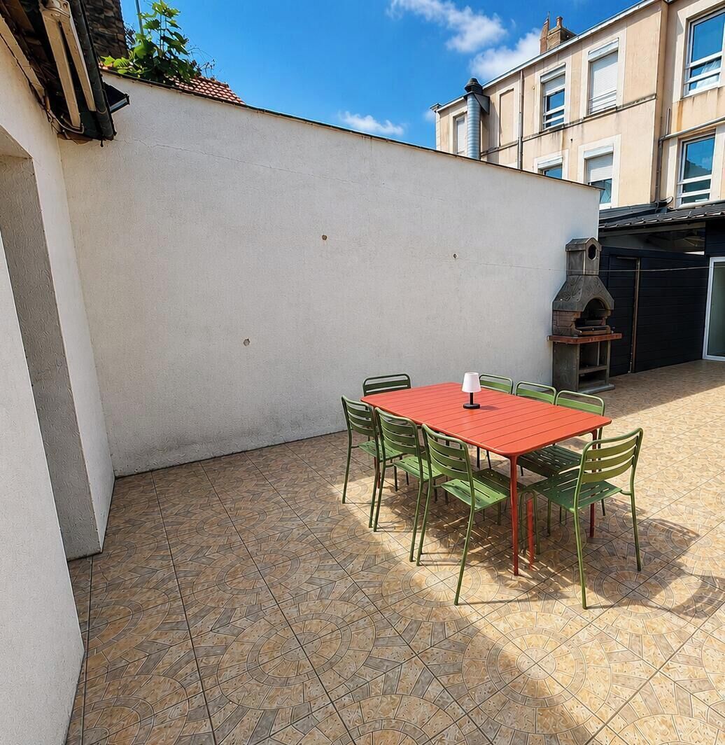 Een terras met een rode tafel en groene stoelen, een barbecue en een witte muur.