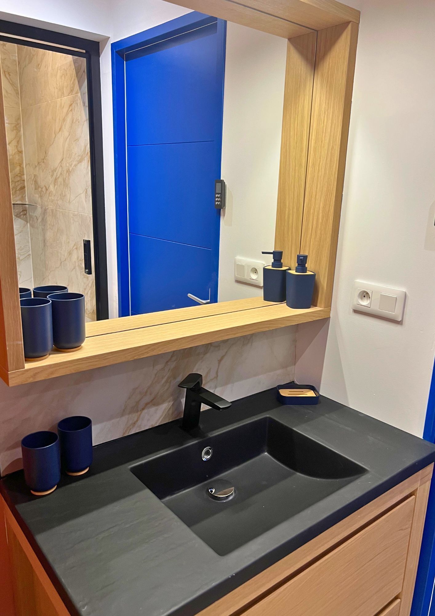 A bathroom with a black sink, blue cups, a wooden shelf, and a blue door.