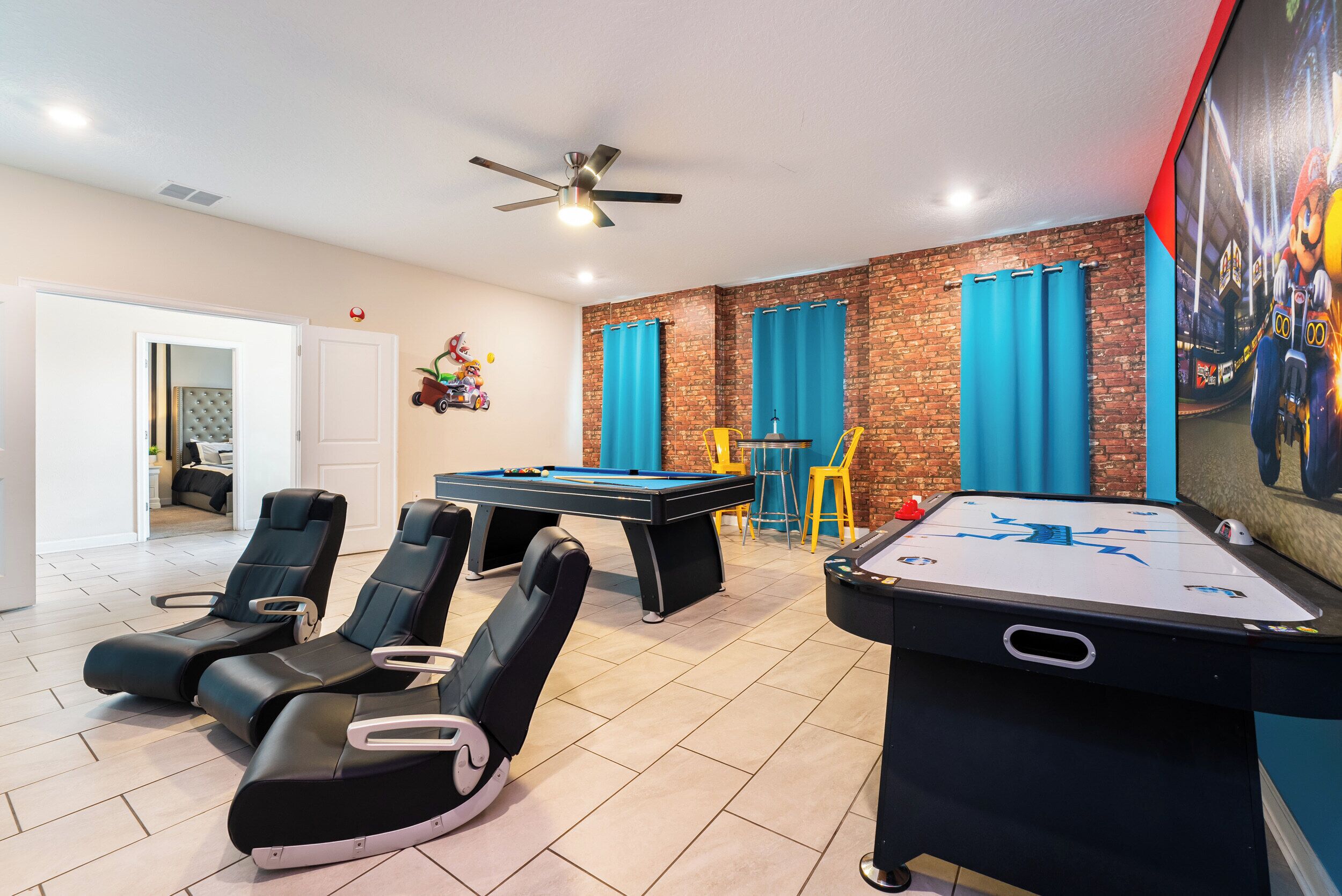 Une table de billard, des fauteuils confortables et un ventilateur de plafond dans une pièce aux murs en briques et aux rideaux bleus.
