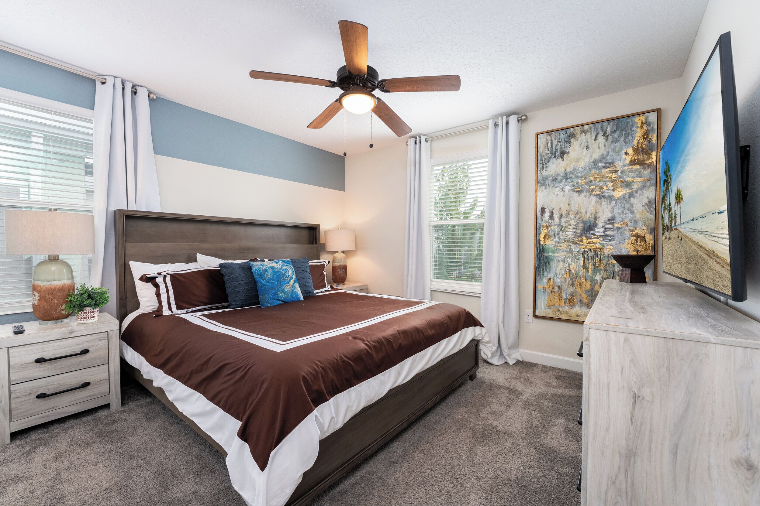 A bedroom with a large bed, a ceiling fan, a nightstand, a lamp, a painting, and a TV.