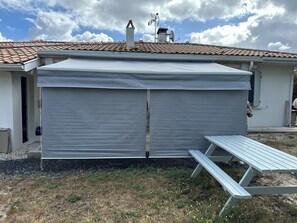 A covered patio area with a table and benches, a white building with a tiled roof, and a clear sky.