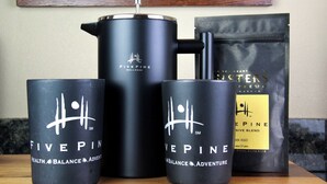 Un ensemble de produits de bien-être Five Pine comprenant une cafetière à piston, deux tasses et un sachet de café en grains.