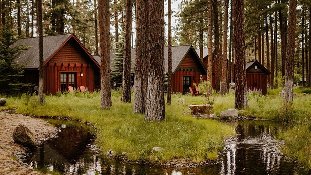 Une rangée de cabanes rustiques, entourées de grands arbres et bordée d’un petit ruisseau.