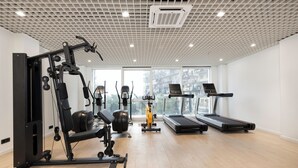 A gym with cardio machines, weight equipment, and a large window offering a city view. - Next White Aparthotel Official (Batumi)