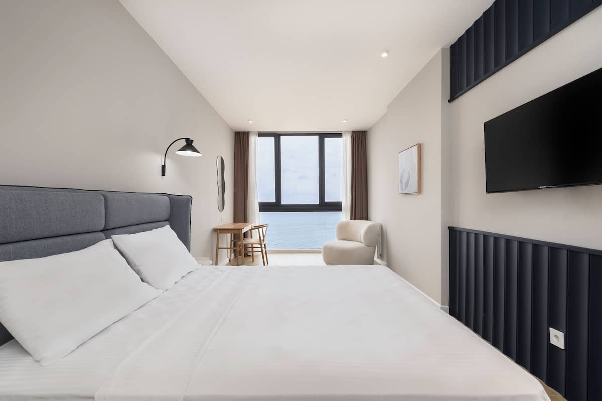 A modern hotel room with a large bed, a desk, a chair, a TV, and a view of the sea.