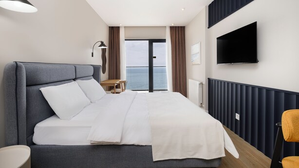 A modern hotel room with a large bed, a desk, a chair, a TV, and a view of the sea through a glass door. - Next White Aparthotel Official (Batumi)