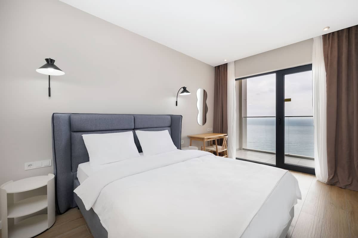 A modern hotel room with a large bed, a bedside table, a small desk, and a view of the sea through a large window.