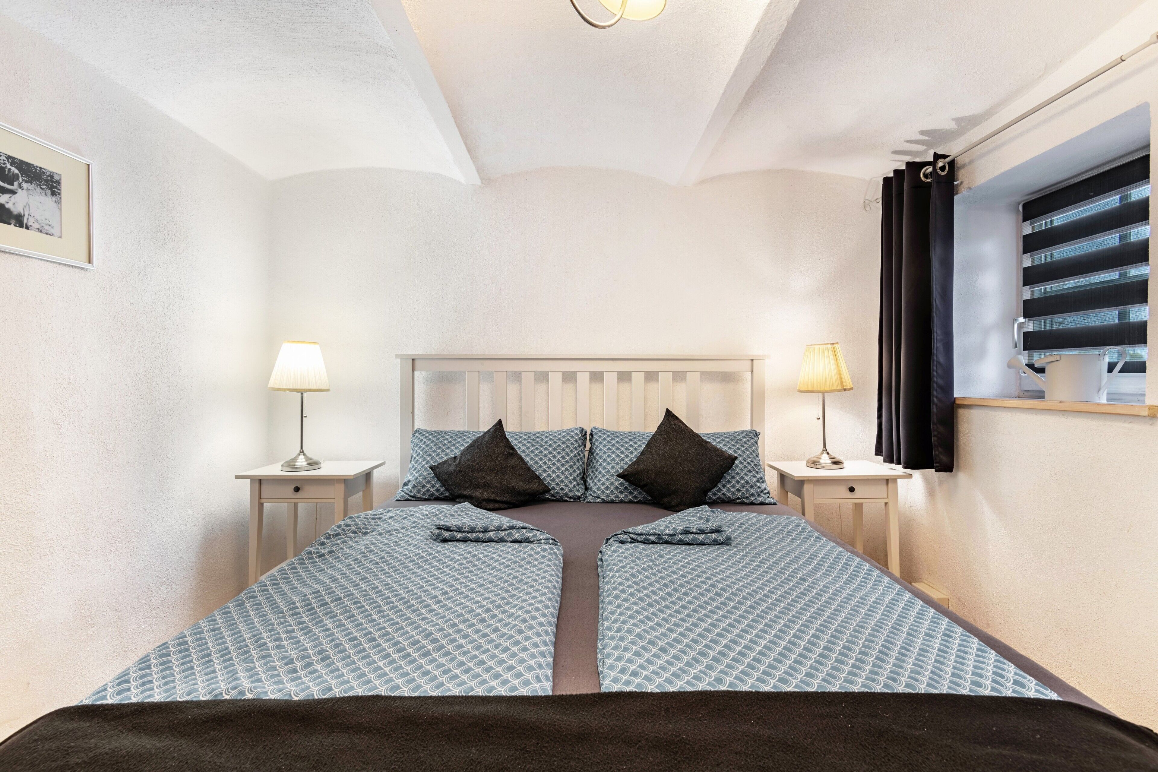 Une chambre à coucher avec un lit placé au centre, entre deux tables de chevet, une fenêtre avec des stores et un tableau encadré accroché au mur.