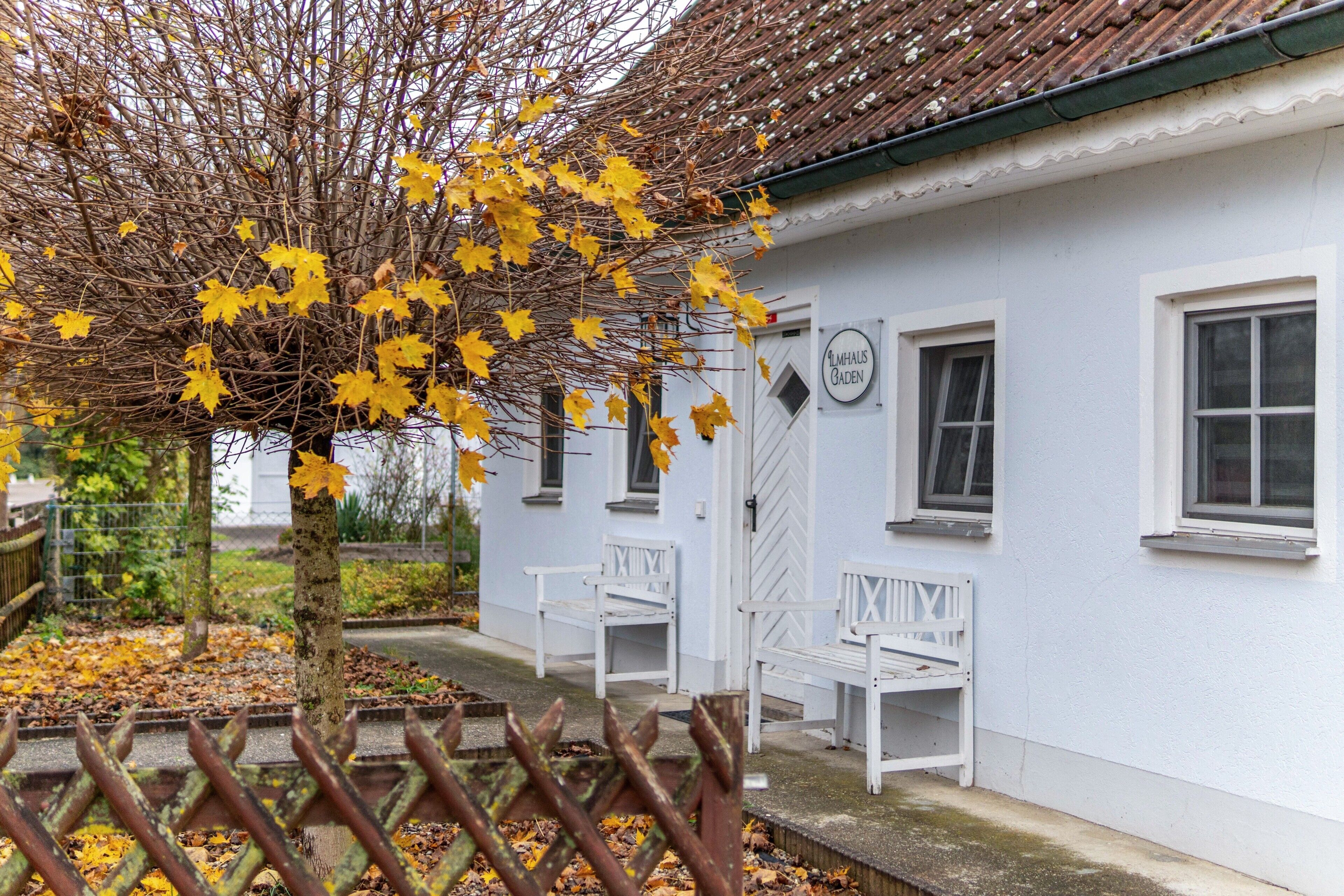 Ein weißes Gebäude mit zwei weißen Bänken davor, ein Baum mit gelben Blättern und ein Schild mit der Aufschrift 'MHAUS GÄDEN'.