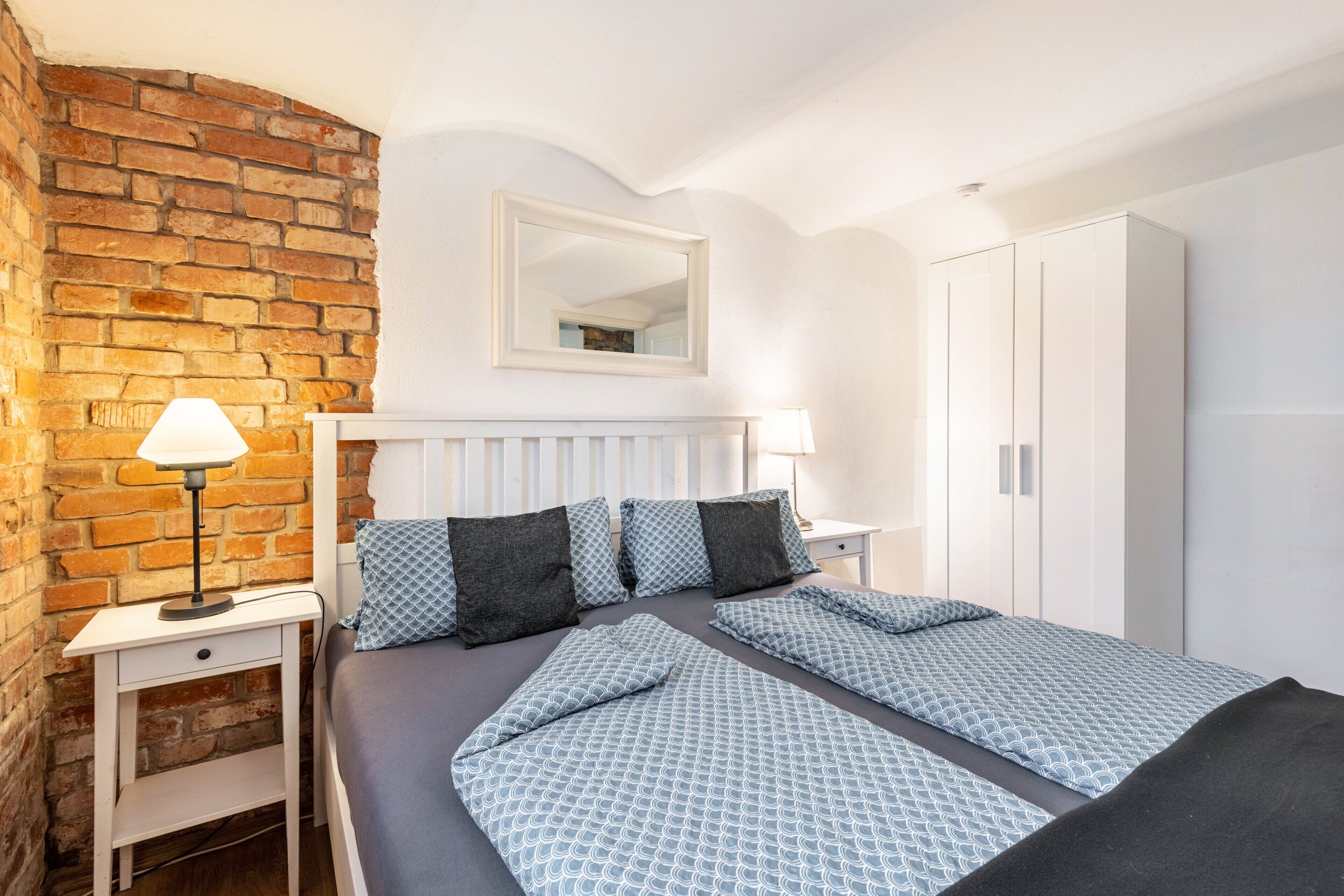 Ein Hotelzimmer mit Backsteinwand, zwei Betten mit gemusterten Bettbezügen, einem weißen Kleiderschrank und einem Spiegel über dem Bett.