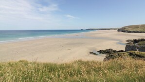 A wide sandy beach with gentle waves, grassy dunes, and rocky outcrops. - Tregundy Farmhouse (Perranporth)