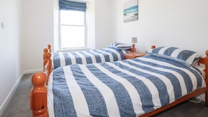 A bedroom with two beds, a window with blue curtains, a bedside table with a lamp, and a painting on the wall. - Tregundy Farmhouse (Perranporth)