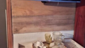 A falra szerelt televízió egy fa szekrény fölött található, a szekrény tetején pedig egy tálca áll, rajta egy pohárral, egy csészével és egy kis tálkával.