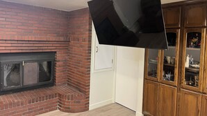 A living room with a brick fireplace, a flat-screen TV mounted on the wall, and a wooden cabinet with glass doors.