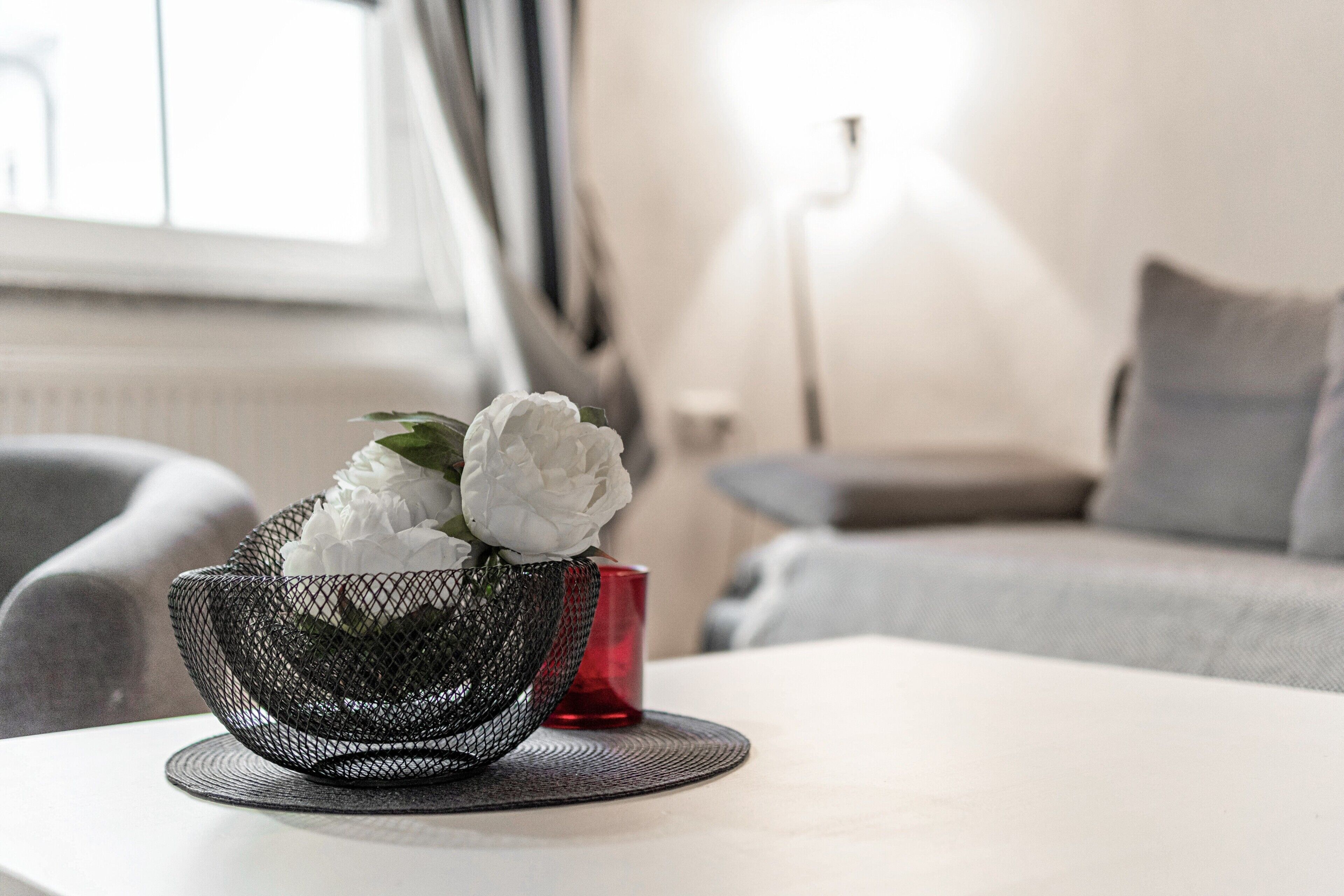 Una sala de estar moderna con una mesa blanca, una cesta de alambre negra con flores blancas, una vela roja y un sofá gris.