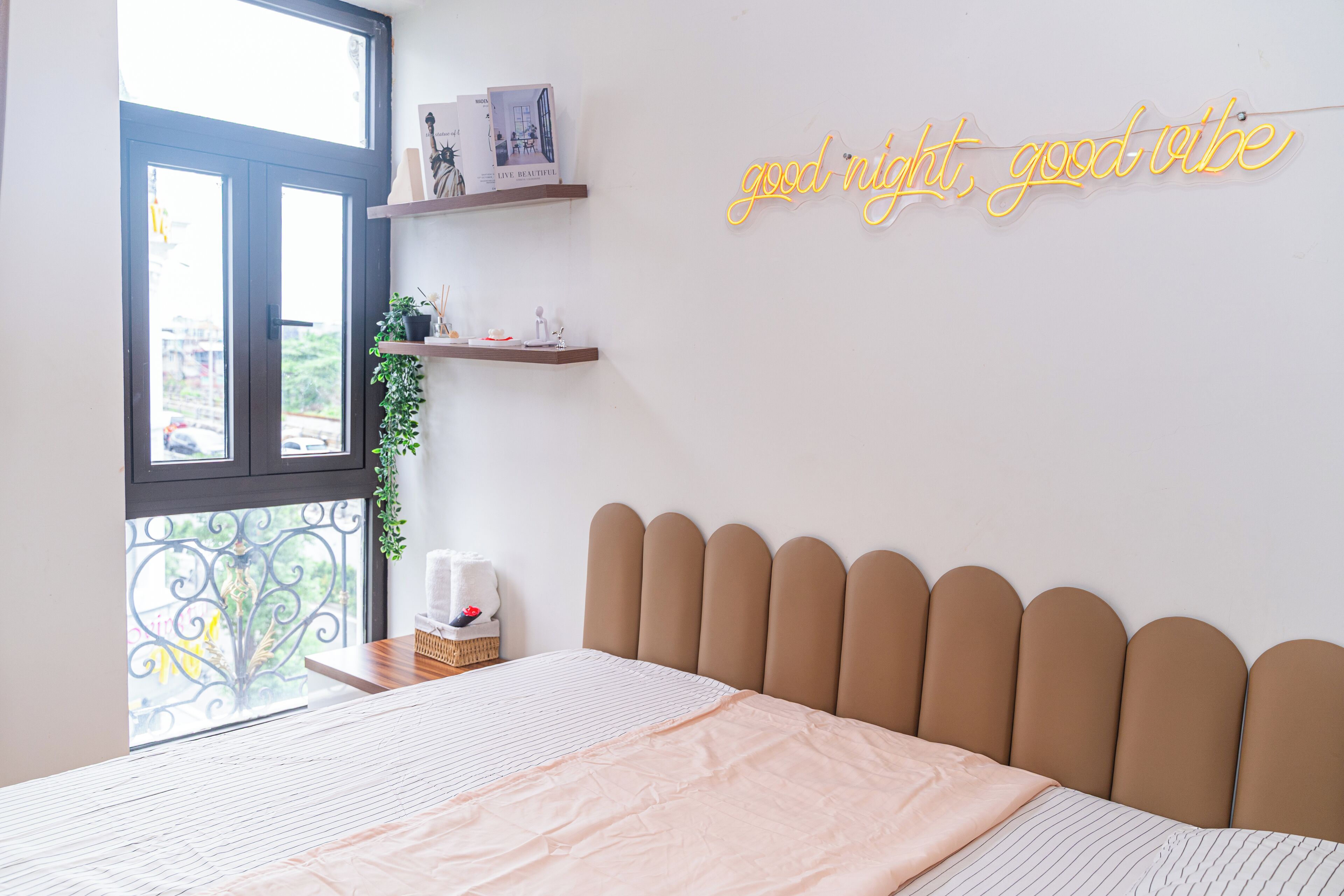A bedroom with a bed, a window, a neon sign, and a shelf with books and a plant.
