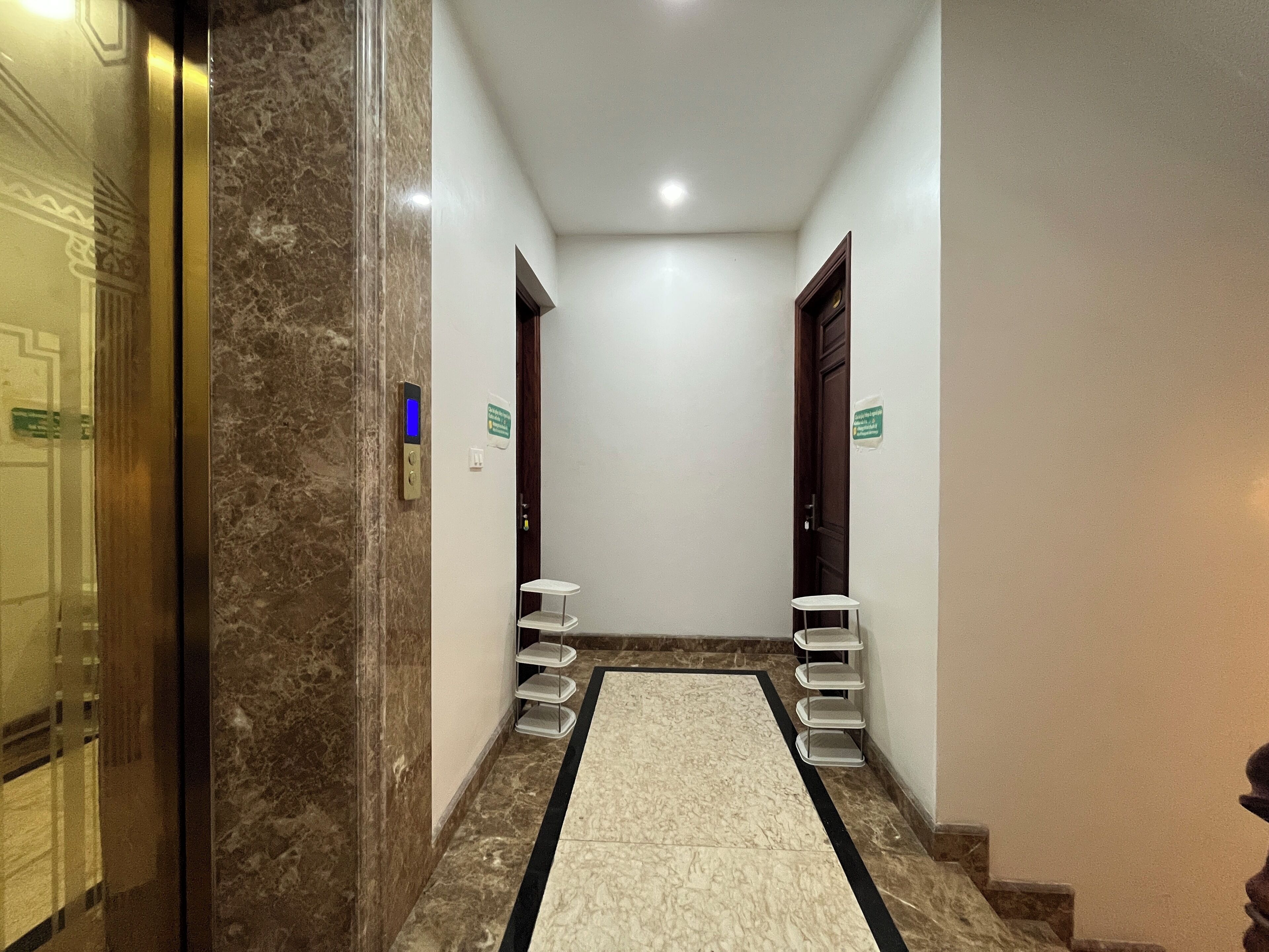 Un corridoio d'hotel con pareti in marmo, pavimento bianco e tappeto centrale bianco. Da un lato e dall'altro, due porte, ognuna con una piccola mensola sottostante.