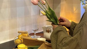 A person holding pink tulips at a table with a glass, a pitcher, and a basket of yellow tulips. - Katka Riverside Homestay Hai Phong (Hai Phong)