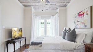 A bedroom with a bed, a TV, a painting, and a window with curtains.