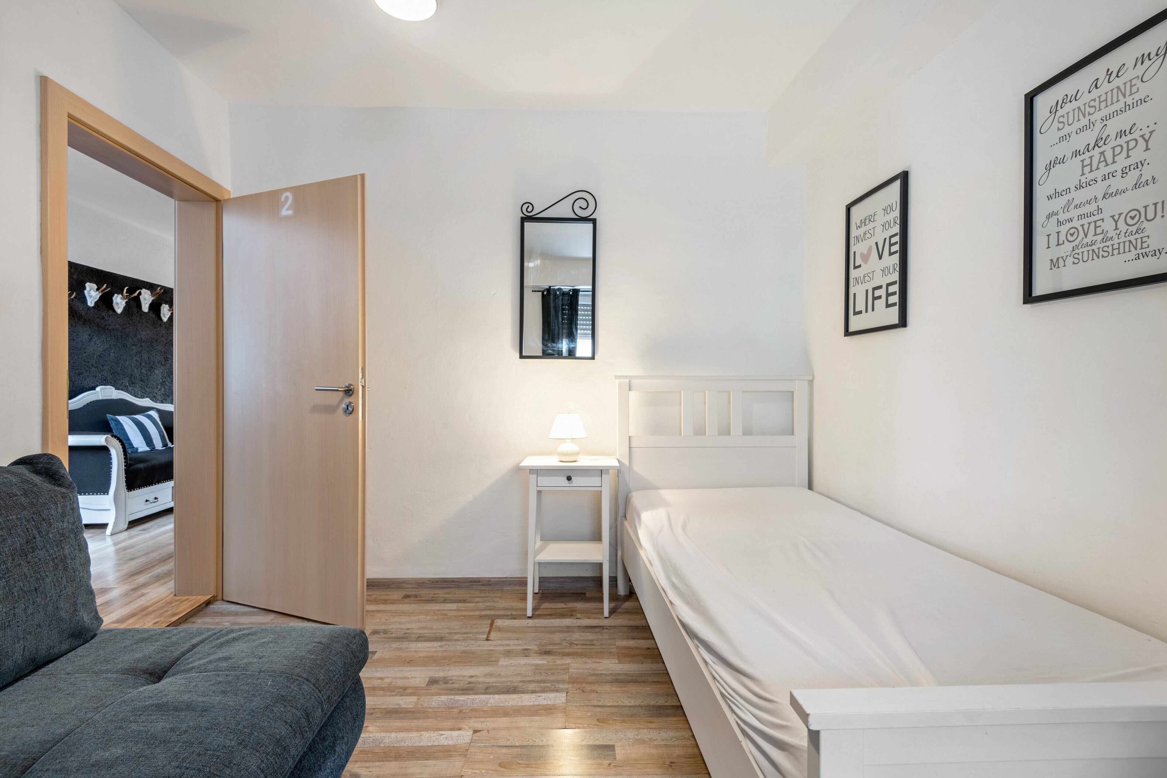 Een slaapkamer met een bed, een nachtkastje met een lamp, een deur en een muur met ingelijste inspirerende citaten.