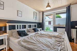Une chambre à coucher avec un lit, des tables de chevet, une fenêtre avec des rideaux et des œuvres d’art murales.