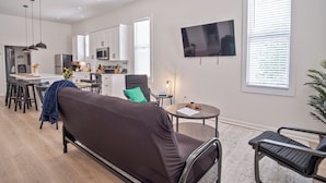 Ein modernes Wohnzimmer mit einer Couch, einem Essbereich mit Stühlen, einer Küche mit Schränken und Geräten sowie einem an der Wand befestigten Fernseher.