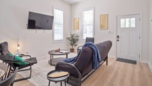 Ein modernes Wohnzimmer mit Sofa, Couchtisch und Sesseln, einem Wand-Fernseher und großen Fenstern mit Jalousien.