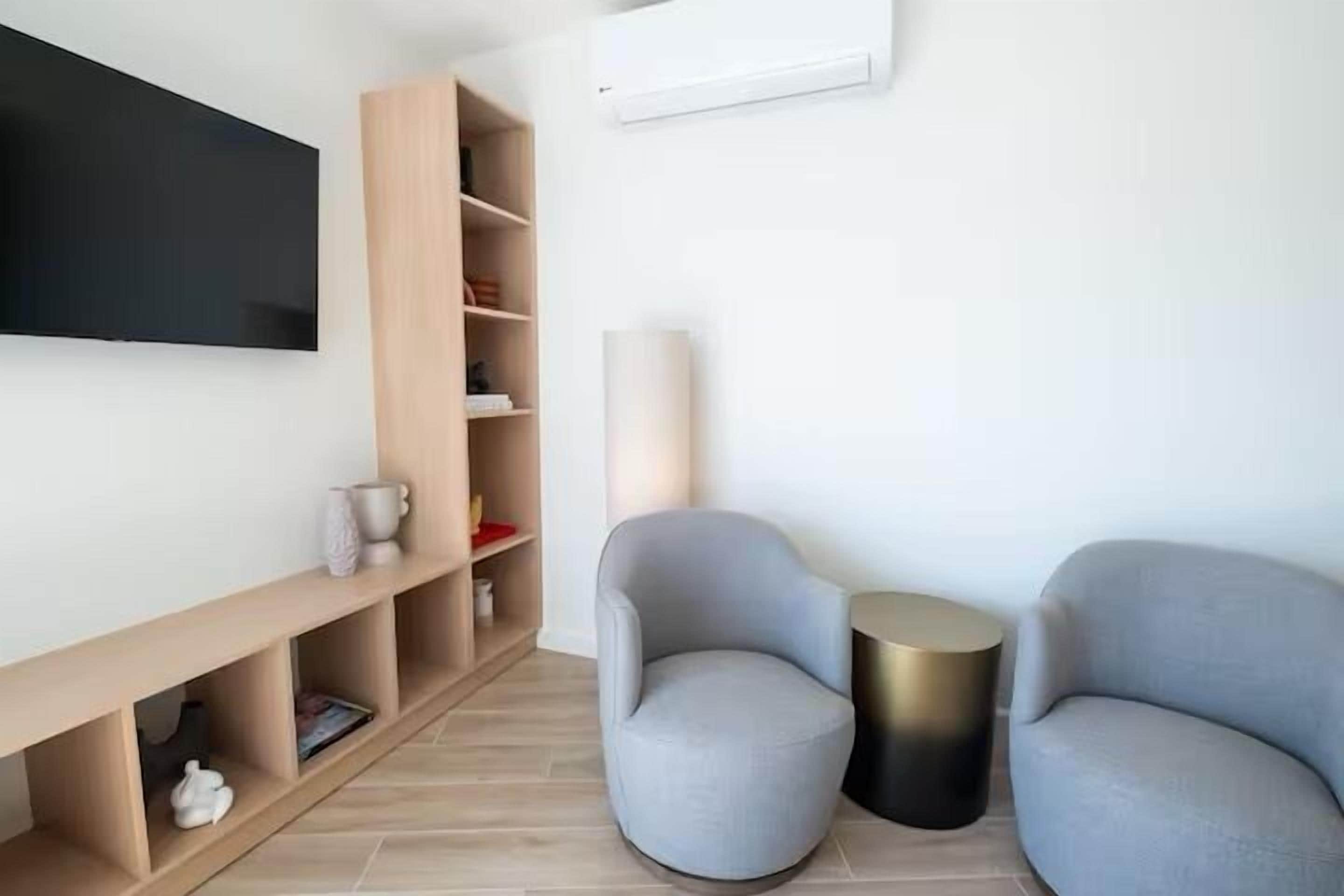 Ein modernes Wohnzimmer mit einem Flachbildfernseher, einer Holzregaleinheit, zwei grauen Sesseln, einem runden Beistelltisch in Goldfarbe und einer weißen Klimaanlage an der Wand.