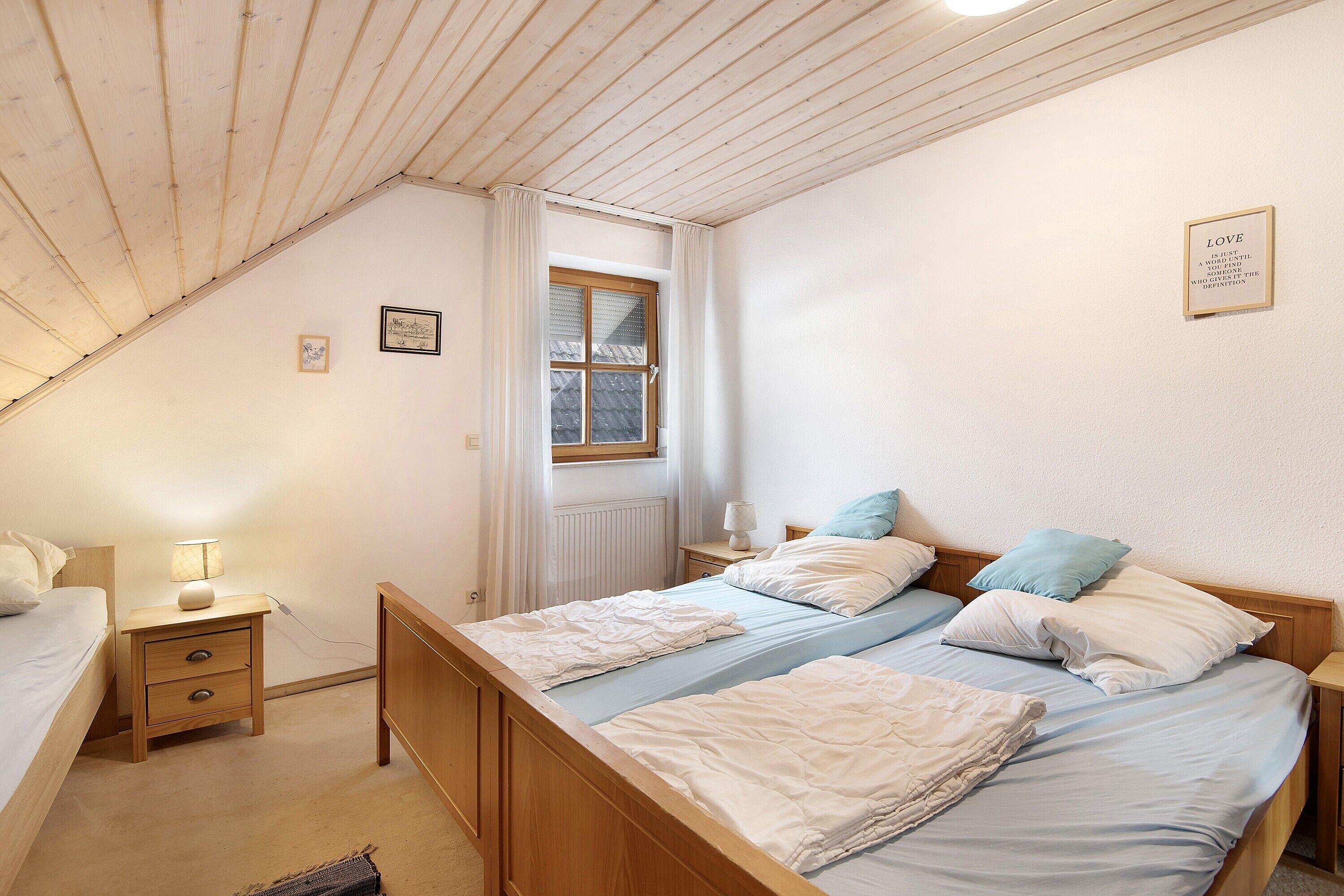 A bedroom with two beds, a window, a bedside table with a lamp, and a framed quote on the wall.