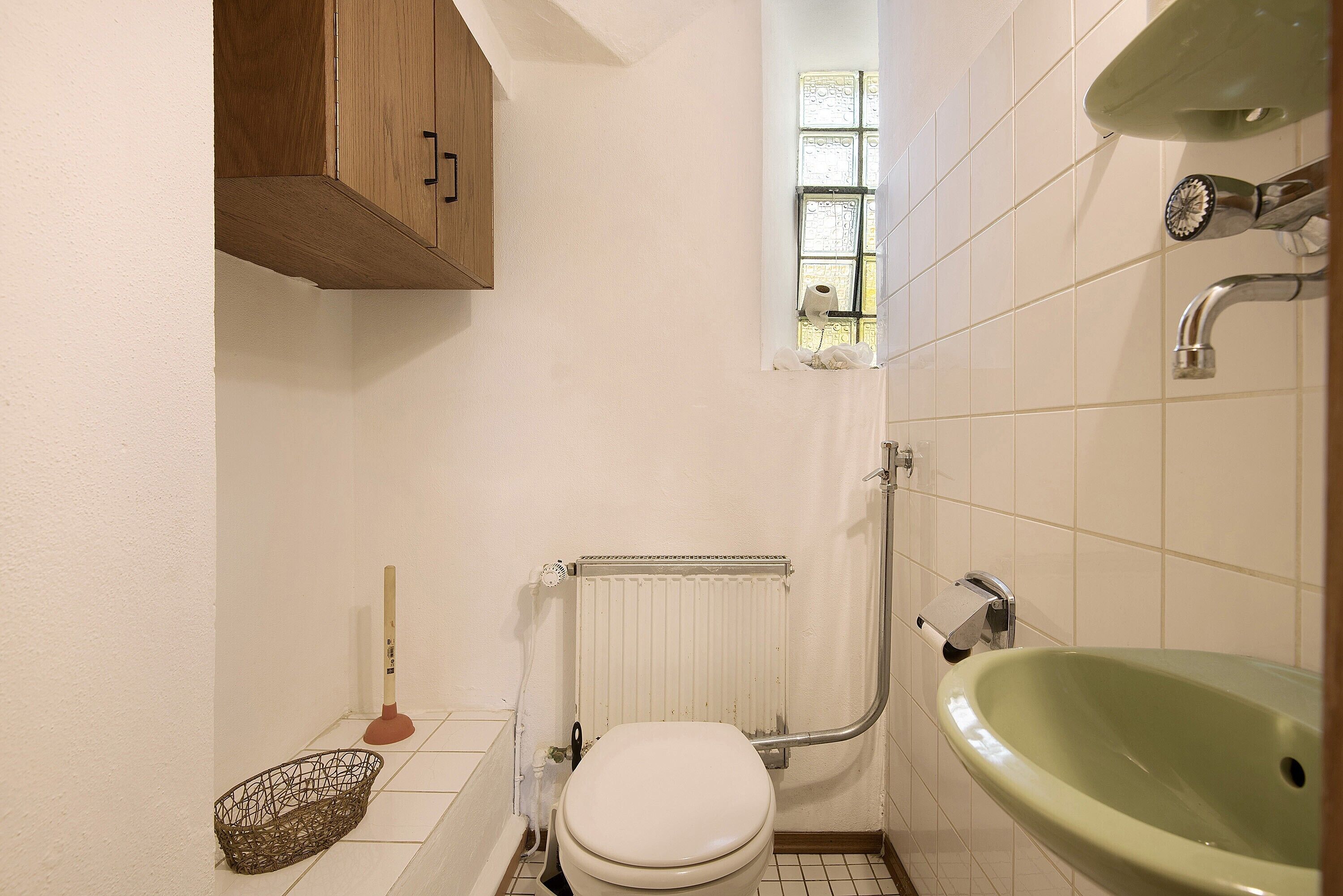 A small bathroom with a toilet, a green sink, a wooden cabinet, a window, and a radiator.