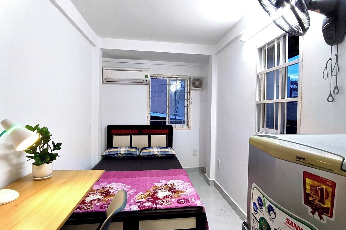 A small, compact room with a bed, a desk, a chair, a radiator, and a window with a view of buildings outside.