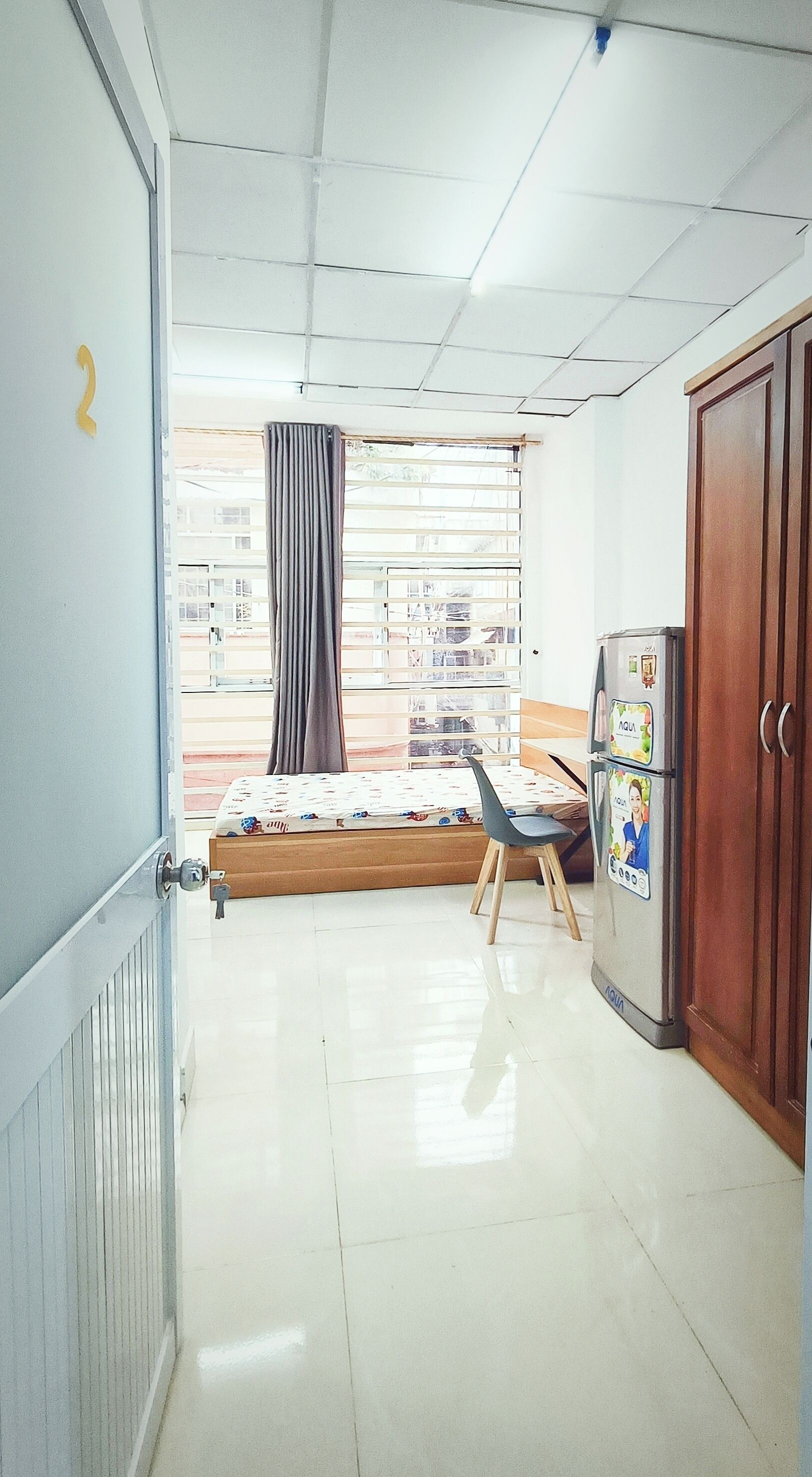 A room with a bed, a chair, a refrigerator, and a window with curtains.