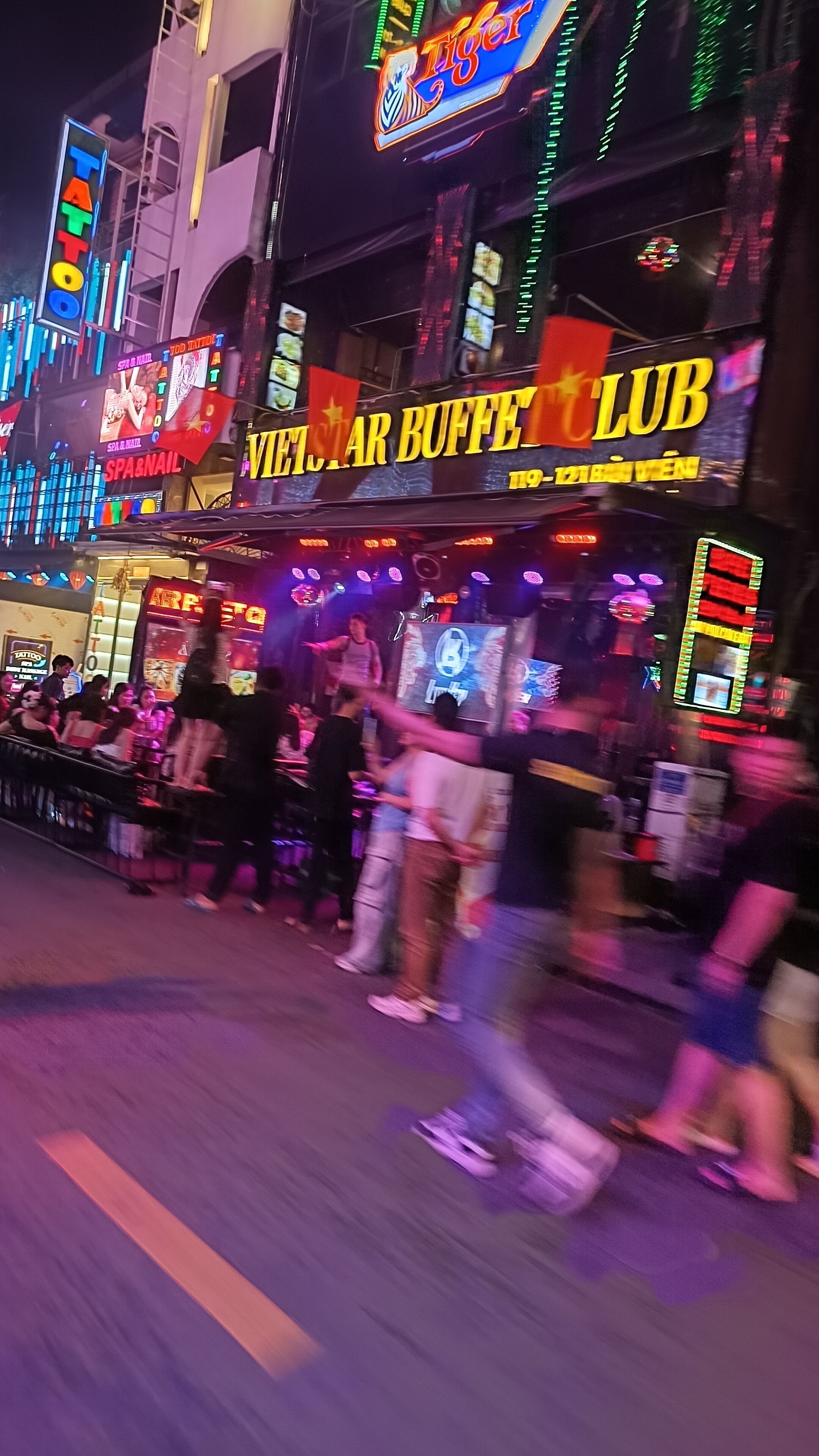 A bustling street scene with neon signs, including "Tiger" and "Vietnam Buffet Club."