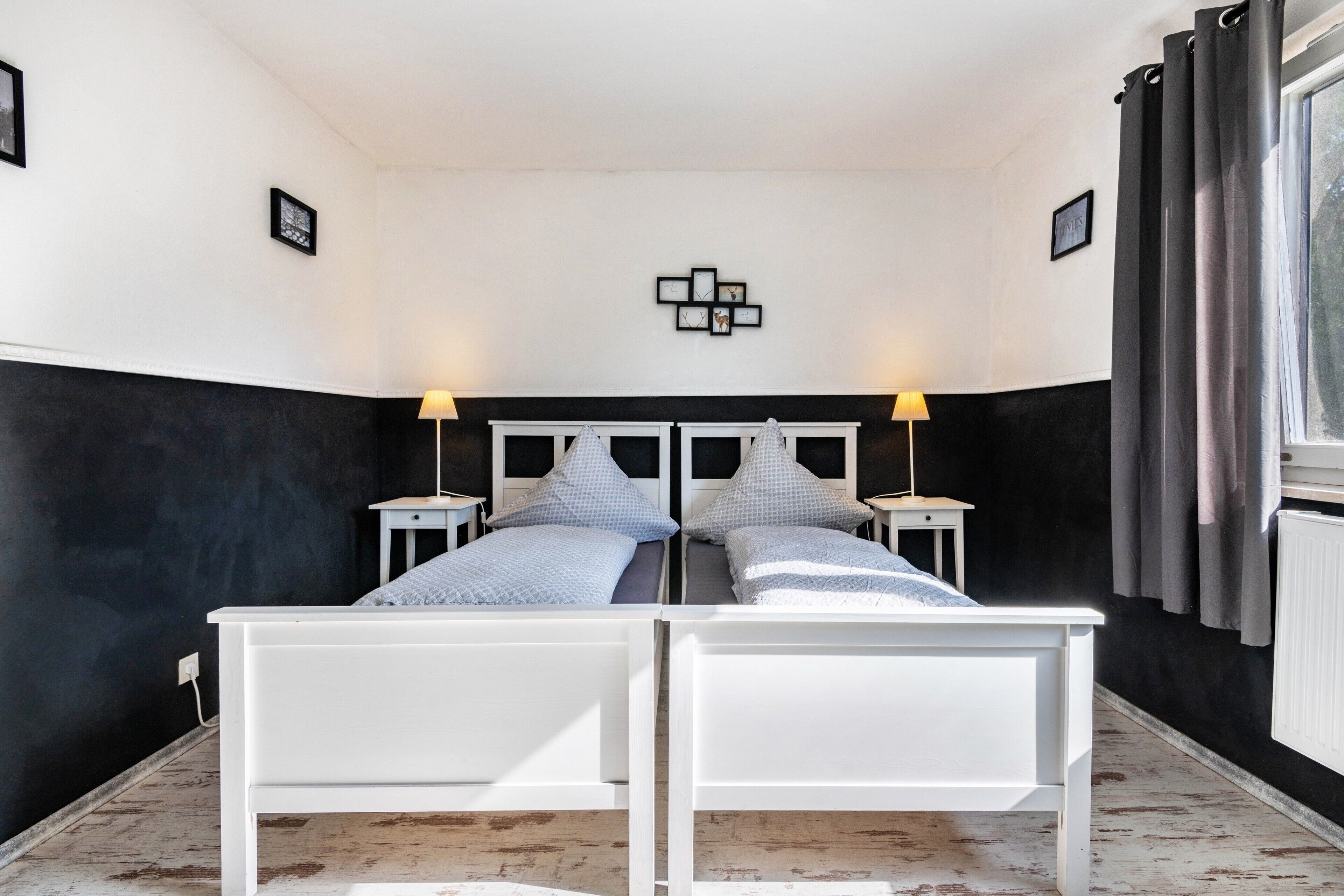 A double bed with white headboards, two bedside tables with lamps, a black and white wall design, and a window with curtains.