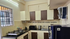 Eine Küche mit Gasherd, Mikrowelle und Kühlschrank. Es gibt Schränke und ein Fenster mit Gitterrost.