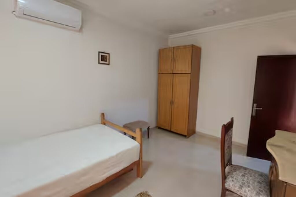A small, sparsely furnished room with a single bed, a wooden wardrobe, a chair, and a small table.